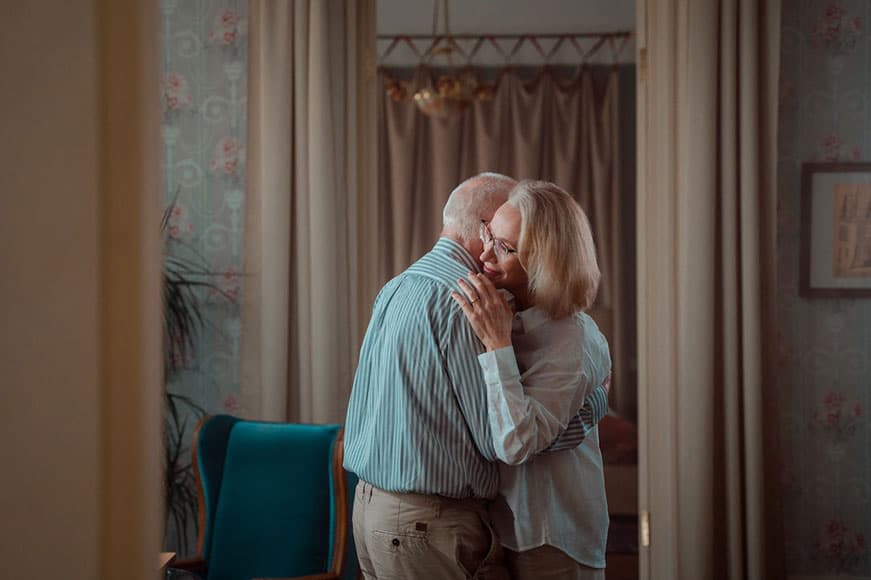 Two seniors in a living room. They’re both standing and hugging one another