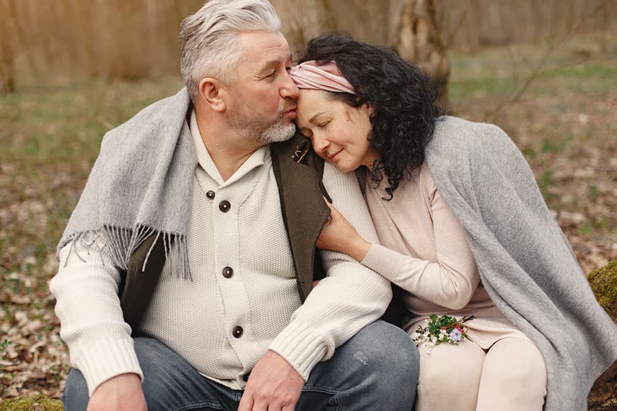 Two people are sitting in a fall park. The lady with shoulder-length dark curly hair has her head lovingly resting on the man with the grey hair's shoulder