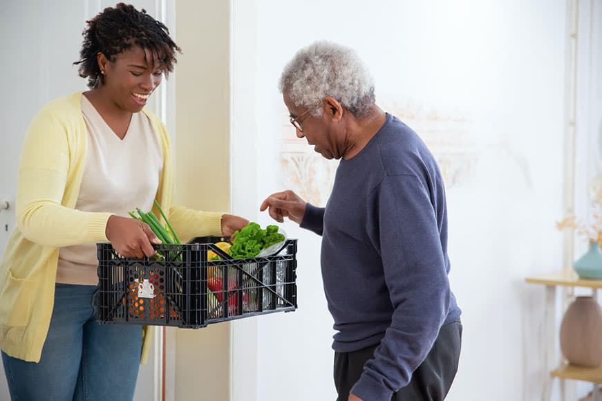 A young woman in a yellow cardigan is holding up a black basket full of produce. A senior man is looking in the basket, picking a piece of produce