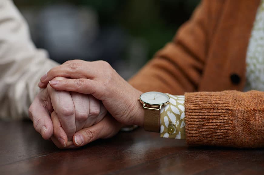 An up-close photo of two seniors holding hands. Their hands are atop a wooden table