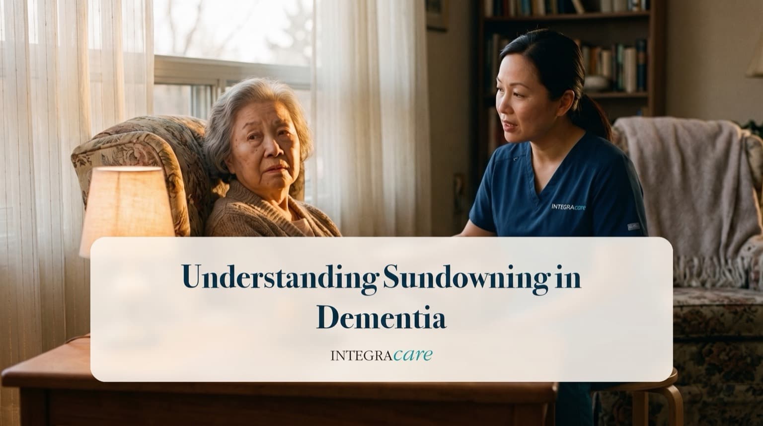 A senior woman sits in an armchair by a bright window while a caregiver speaks with her