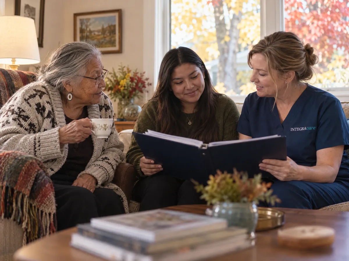 A nurse in navy scrubs and a young woman go through a binder together on the sofa while the young woman's grandmother, teacup in hand, joins in from an armchair beside them, autumn leaves filling the window.