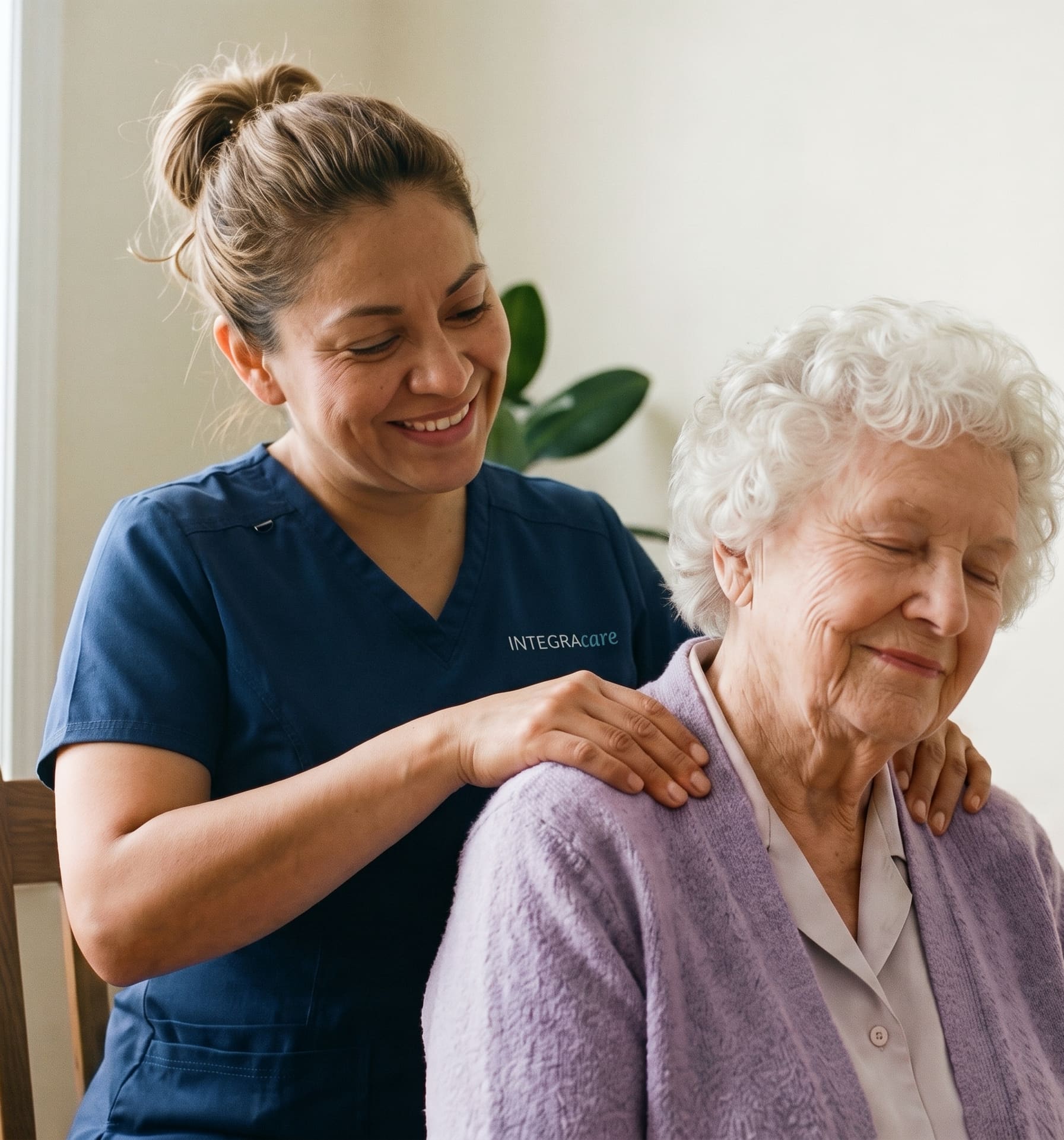 A smiling massage therapist in navy scrubs rests both hands on the shoulders of a seated older woman in a lilac cardigan, whose eyes are closed.
