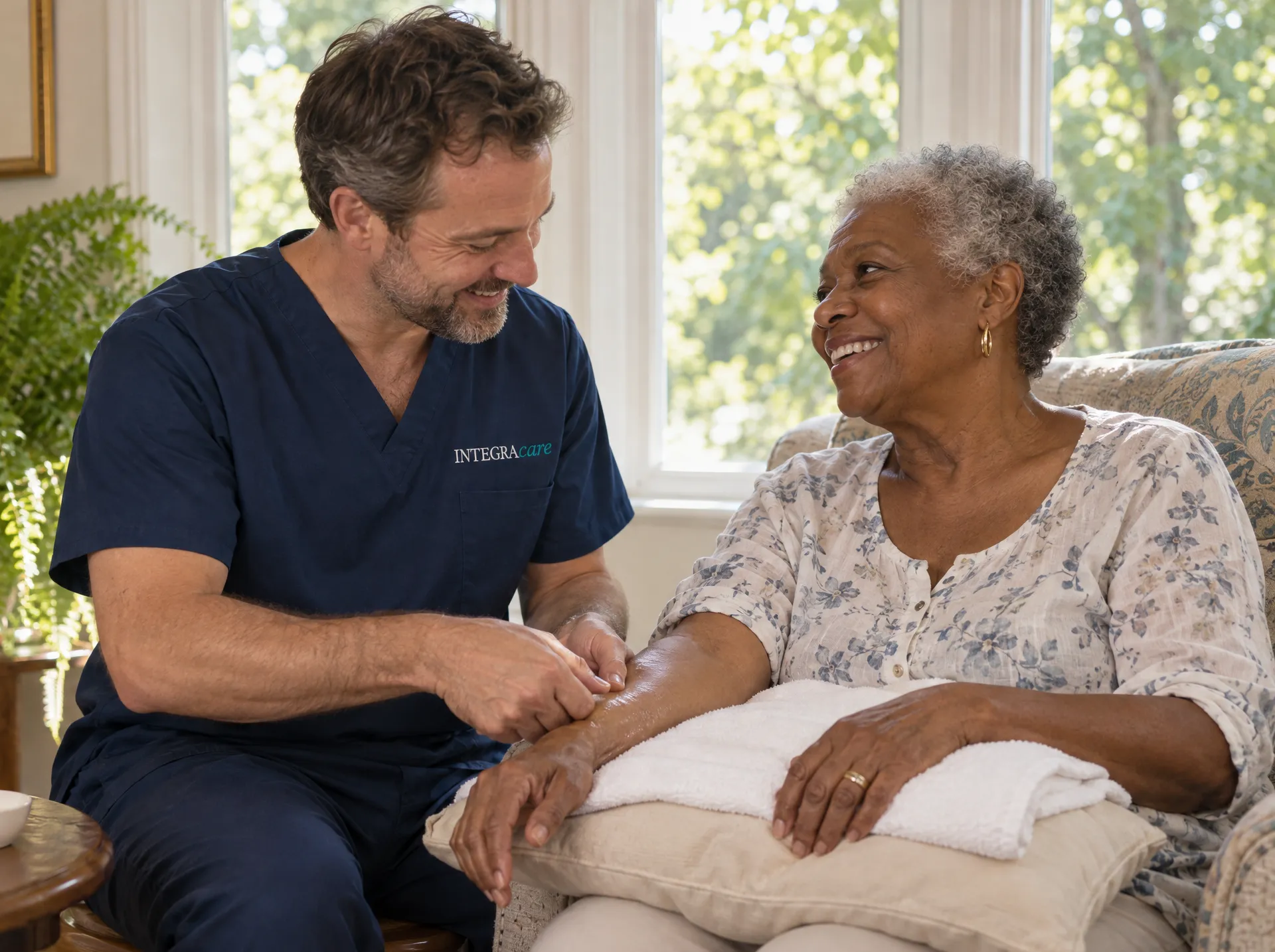 A massage therapist in navy scrubs works along the forearm of a smiling older woman in an armchair, her arm resting on a folded towel over a pillow, in a sunny room with leafy windows behind.