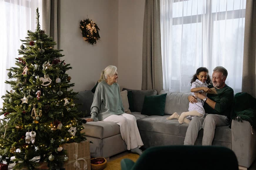 A senior lady sitting on a couch by a Christmas tree. A senior man sits on the other side with a little girl on his knee