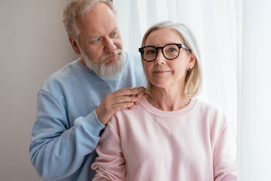 A young senior man in a blue sweater and a woman in a pink sweater facing the camera. He has his hands on her right shoulder.