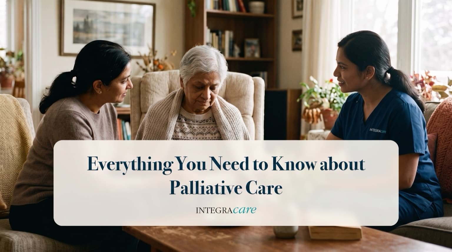 An older woman wrapped in a knitted shawl sits in an armchair at home while a family member and a caregiver in navy scrubs sit on either side, talking with her.