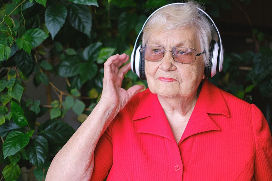 A close-up of a white senior lady. She’s wearing glasses and a red shirt. She has big white headphones on.