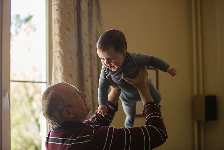 A senior man smiling, holding a laughing baby in the air