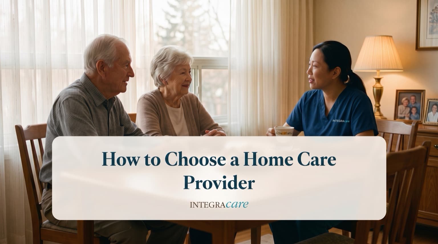 A senior couple sitting at their dining table talking with a caregiver in navy scrubs over a cup of tea