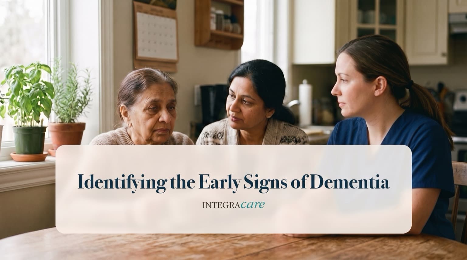 A caregiver in scrubs sits at a kitchen table talking with a senior woman and her adult daughter