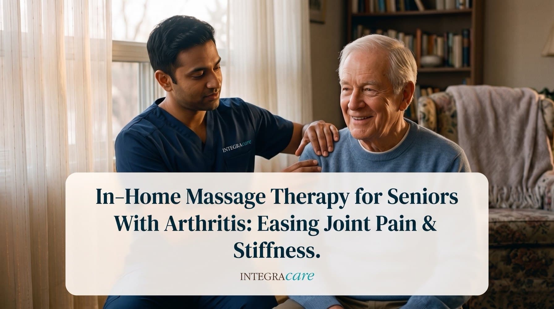 A massage therapist in navy scrubs rests a hand on the shoulder of a smiling white-haired senior man in a blue sweater, seated together in a sunlit living room