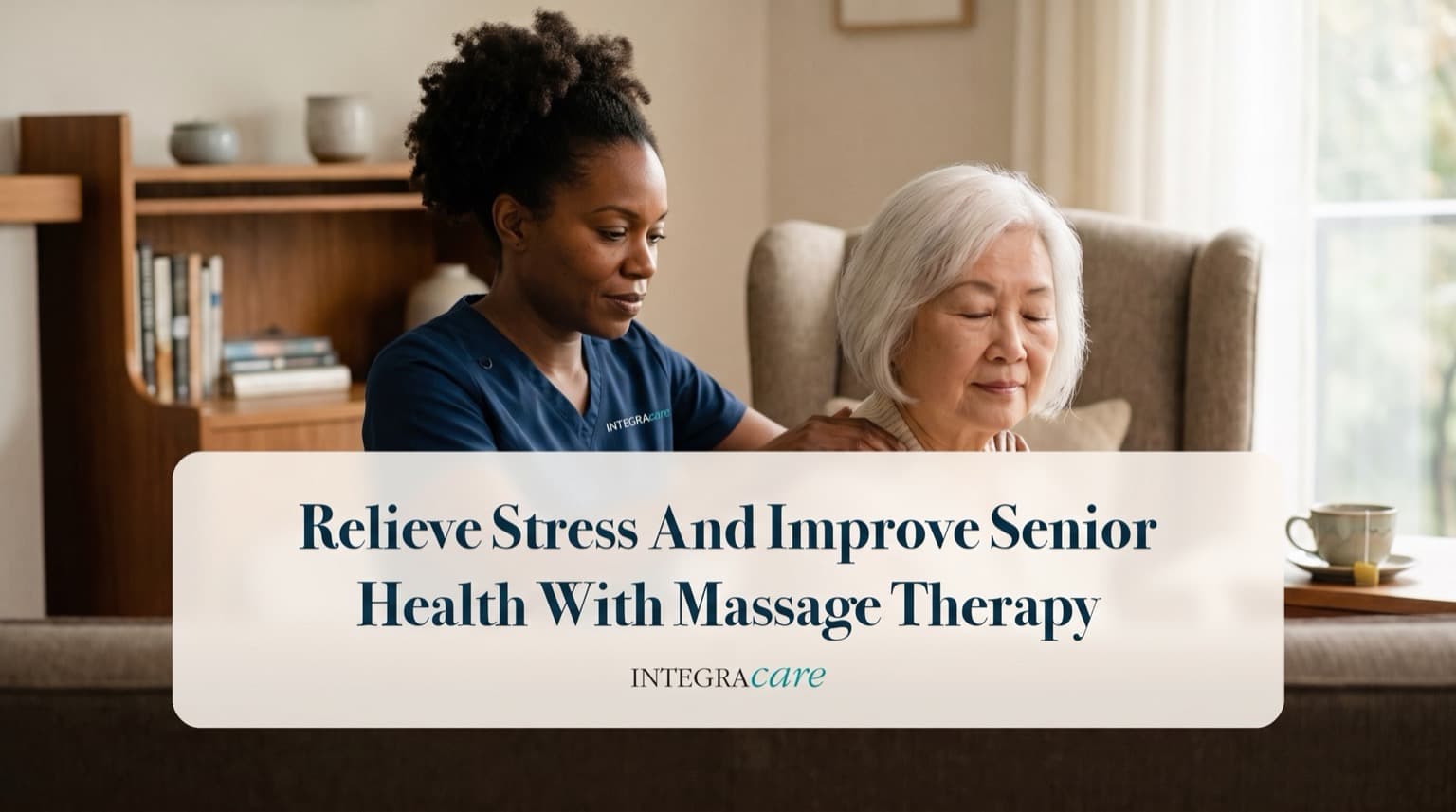 A massage therapist in navy scrubs gently massages the shoulders of a white-haired senior woman seated in an armchair in her living room