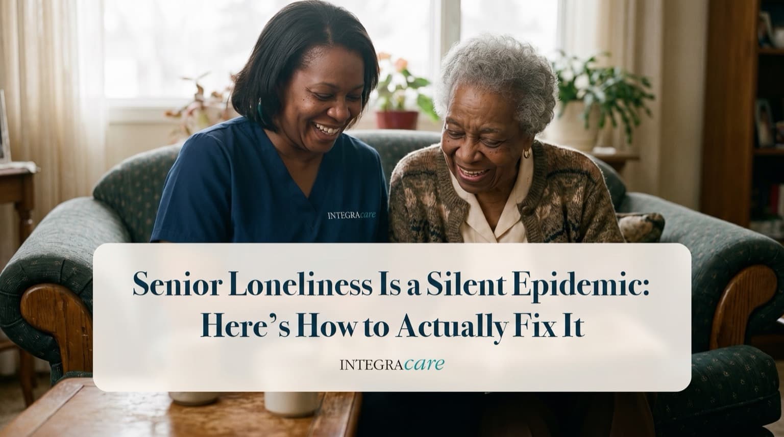 A smiling caregiver in navy scrubs and a senior woman laugh together on a couch in a bright living room, under the title Senior Loneliness Is a Silent Epidemic: Here's How to Actually Fix It