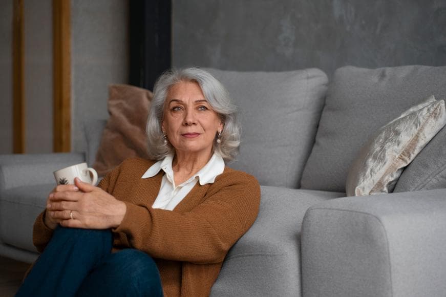 Senior woman sitting on the ground in front of a sofa holding a cup