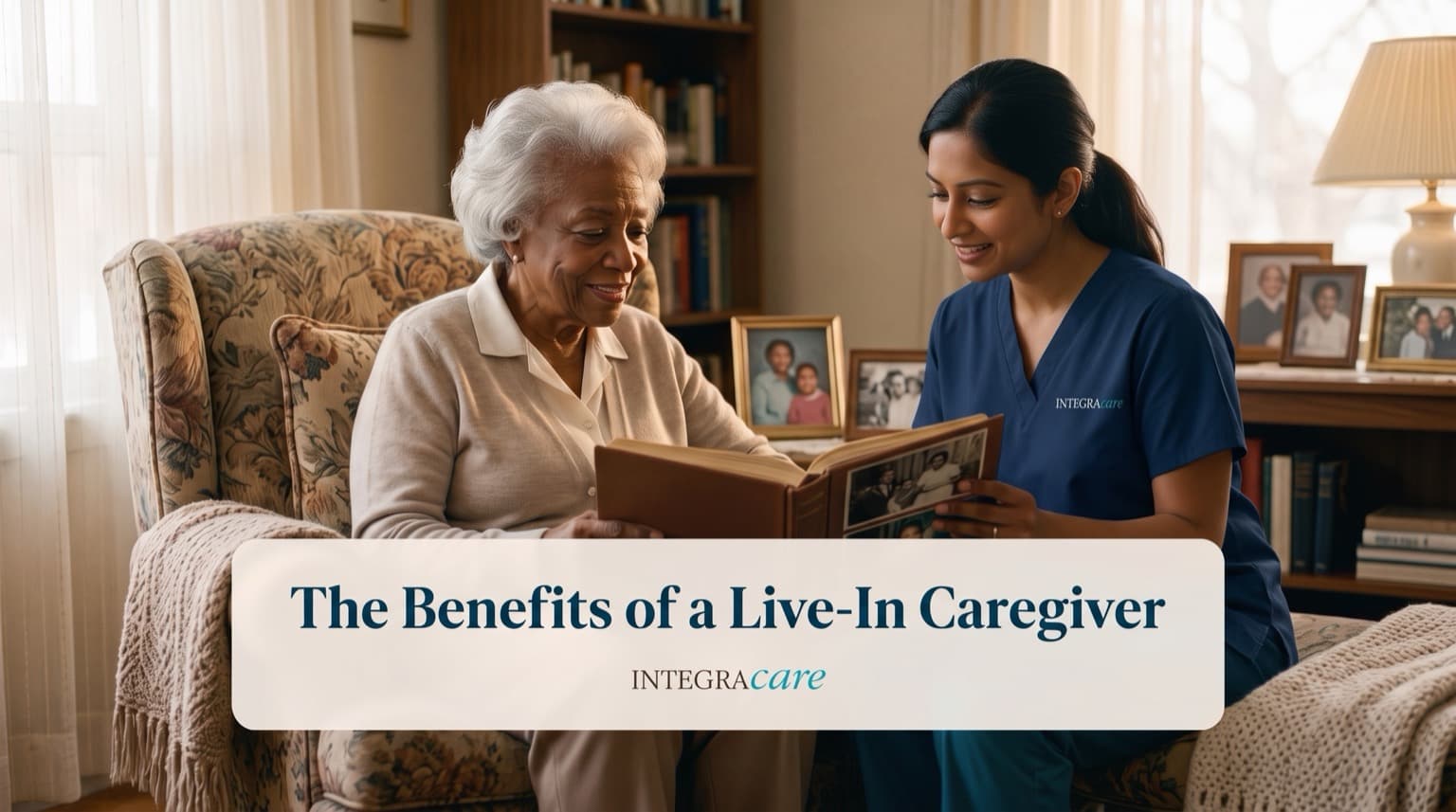 Caregiver in navy scrubs looking through a photo album with a senior woman seated in a floral armchair in her living room
