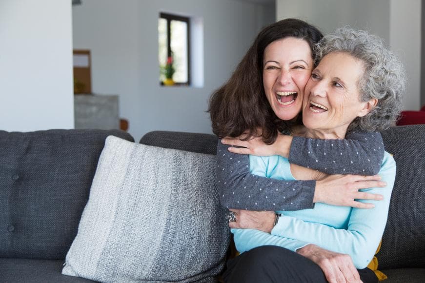 Mother and daughter sitting on couch, daughter hugging mother from behind both smiling