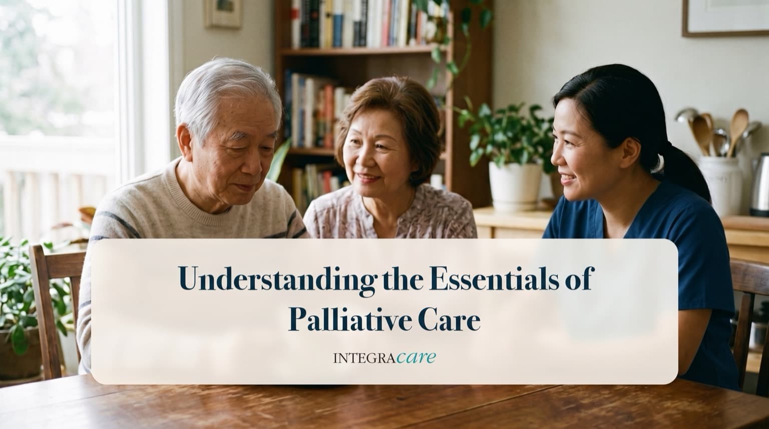 An older couple sits at a wooden dining table in a bright home, talking with a smiling caregiver in navy scrubs, with bookshelves and plants behind them.