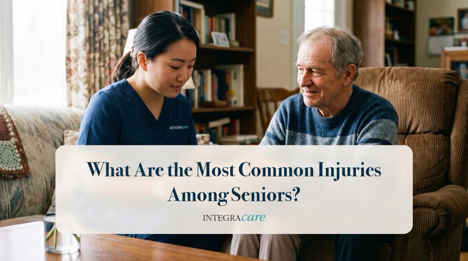 A caregiver in navy scrubs sits at a coffee table with a smiling senior man in his living room, under the title What Are the Most Common Injuries Among Seniors?