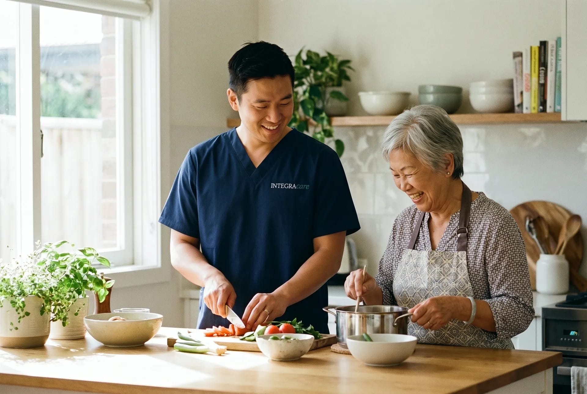 A smiling caregiver in a navy scrub top chops vegetables while an older woman stirs a pot at a sunny kitchen counter.