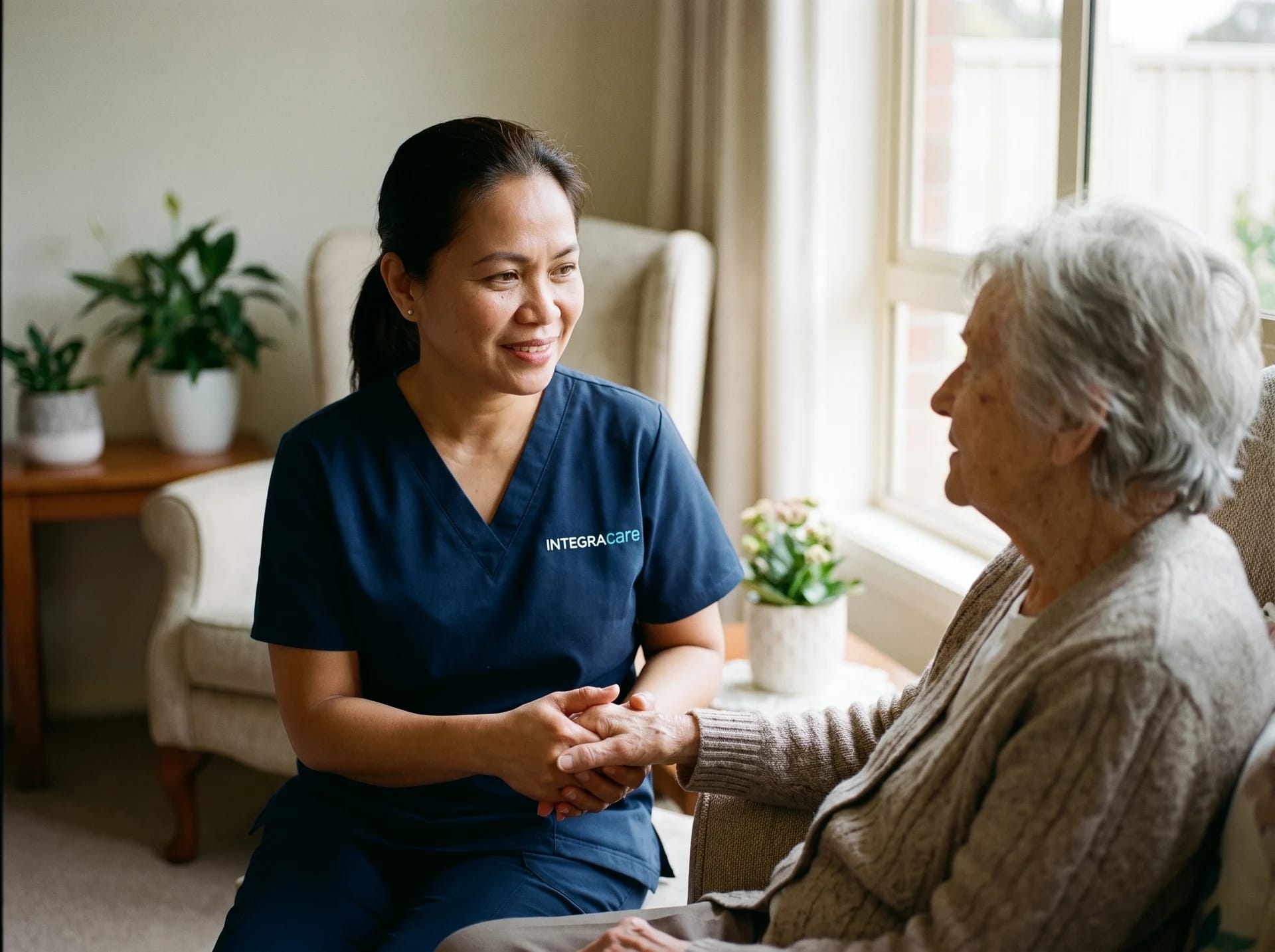 A caregiver and an older woman hold hands and talk quietly beside a bright window.