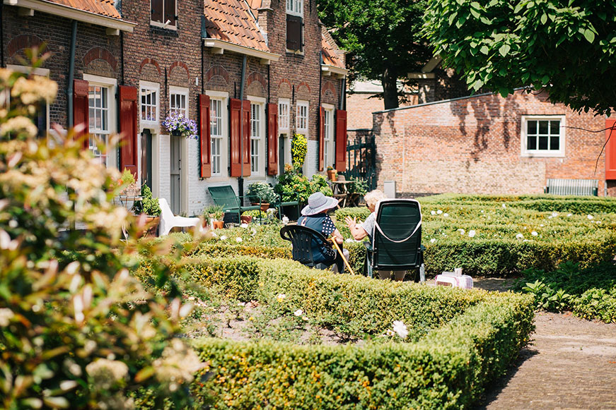Two Seniors sitting at the door of a beautiful red brick building in an immaculately landscaped garden