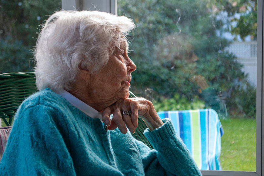A senior lady in a green cardigan. Sitting in a sun house. She has her chin resting on her hand and she’s looking to the side