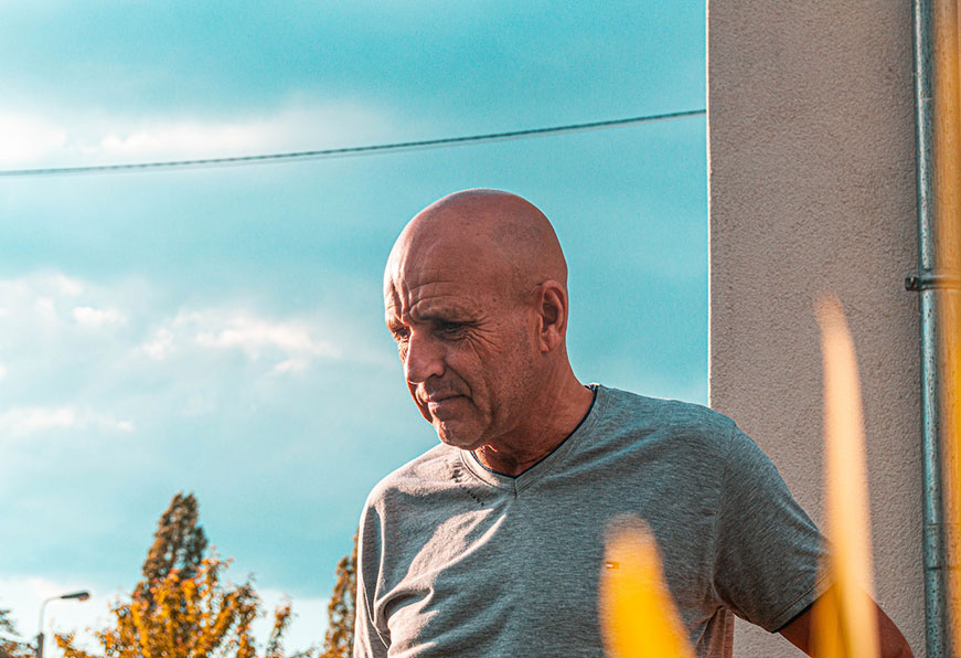 A close-up of a bald man standing outside a building on a sunny day. He is wearing a grey t-shirt and looking at the ground