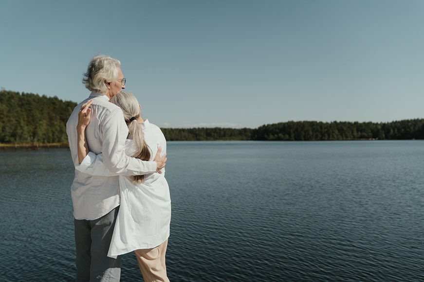 Two seniors embracing, looking out over a blue lake that’s surrounded by trees. They’re both wearing white shirts