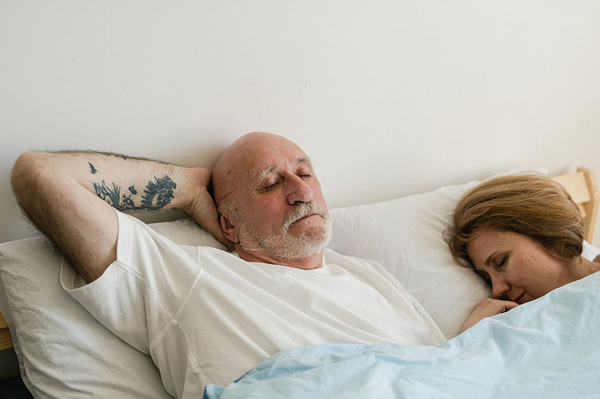 Two seniors lying in bed. The lady is asleep; the man has his right arm behind his head and sits slightly elevated by his pillow