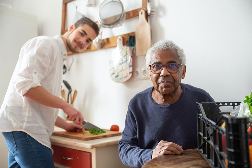 A younger man in the background chopping vegetables. An older man is in the foreground sitting at a wooden table