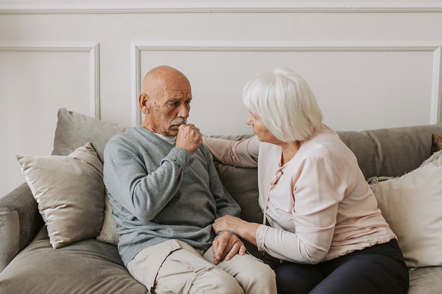 Two seniors sitting on a grey couch. The lady consoles the man; he looks pensive and has his hand to his mouth