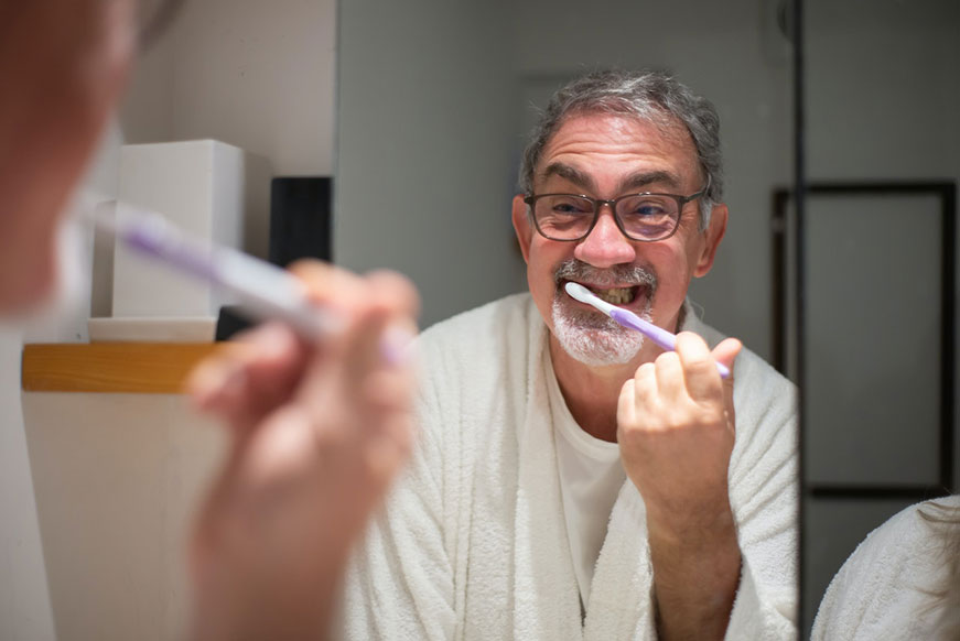 A senior man in a white tee-shirt and black glasses. He's looking in the mirror, brushing his teeth
