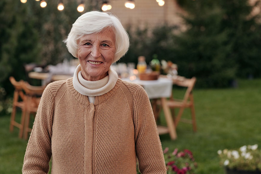 A cheerful senior lady. She’s standing in a green yard. She's wearing a beige sweater with a white roll-neck underneath