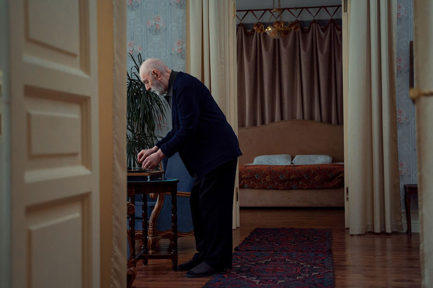 A senior man in a room living room with a bedroom in the background. He’s putting a record on a record player.