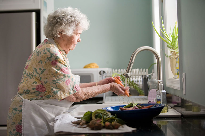 A white senior lady in a pastel green kitchen, she's wearing an apron washing a carrot over a sink