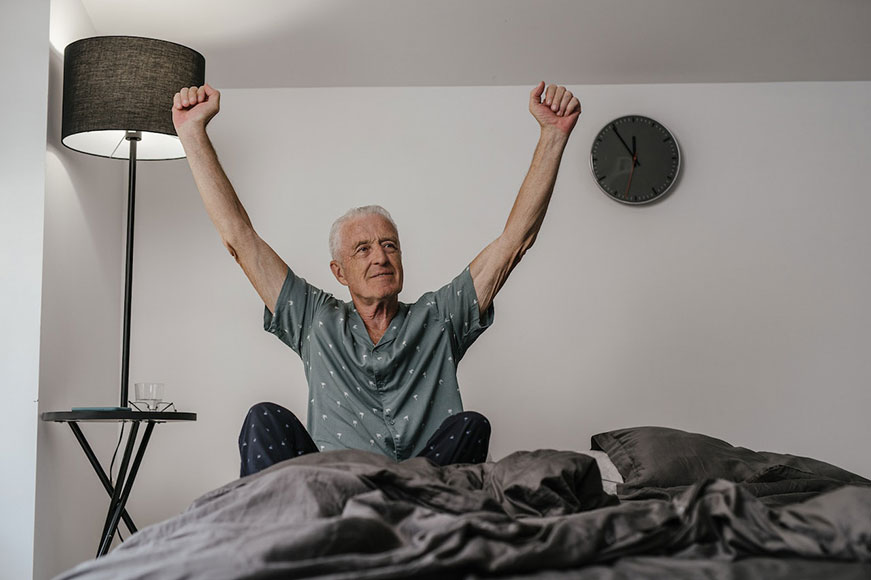 A white senior man in green spotted pjs, stretching in bed
