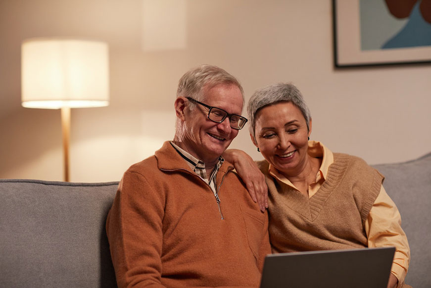 A senior couple on a grey couch watching a laptop and smiling. He wears an orange fleece; she wears a tan sweater vest.