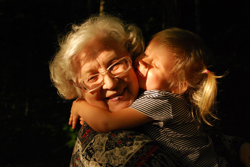 A senior lady holding a little girl in a beam of sunshine. Their faces are close together and they’re both smiling