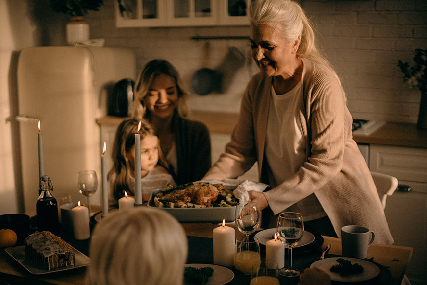A senior lady serving dinner in a large baking dish. A woman sits in the background with a little girl on her knee.