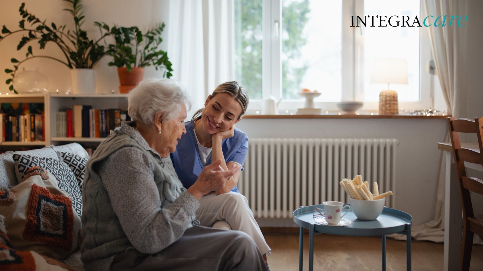 Integracare caregiver sharing a warm conversation with an elderly woman in a cozy living room, symbolizing comfort, dignity, and companionship in home care.